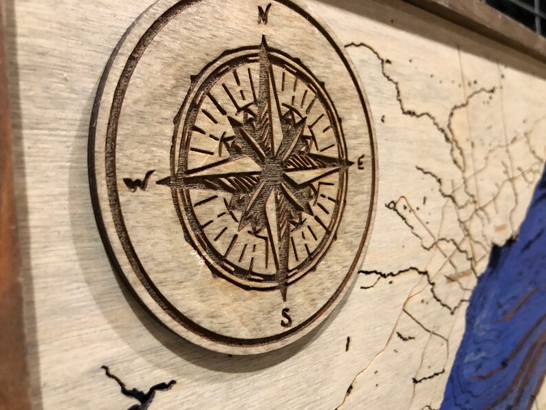 Pu&ograve; includere: Una bussola in legno con un design dettagliato, che presenta le lettere N, S, E e W. La bussola &egrave; posizionata su uno sfondo in legno chiaro con una mappa blu e bianca.