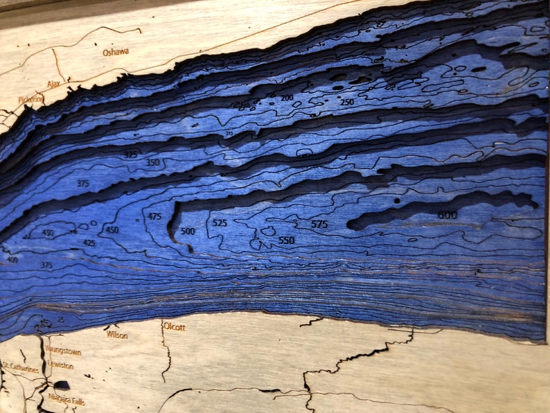 Pu&ograve; includere: Una mappa di legno blu di un lago con curve di livello che indicano la profondit&agrave; in piedi. La mappa mostra la profondit&agrave; del lago che va da 100 a 600 piedi. La mappa include anche i nomi delle citt&agrave; e dei paesi intorno al lago, tra cui Olcott, Wilson, Youngstown, Lewiston e Niagara Falls.