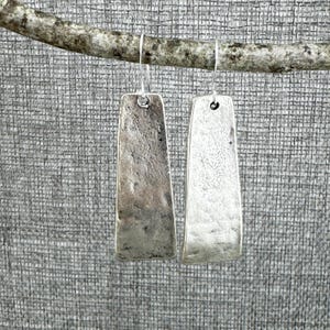 May include: A pair of silver-toned drop earrings featuring elongated, rectangular metal plates with a hammered, textured finish. Each earring hangs from a simple silver wire hook, showcasing a slightly tapered shape and a weathered, metallic appearance.