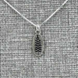 May include: A silver-colored necklace with a small, oval pendant featuring a cut-out tree design. The pendant hangs from a delicate chain, creating a simple and elegant accessory. The necklace is displayed against a textured gray background.