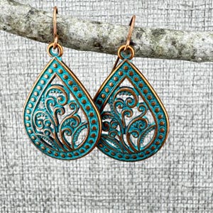 May include: A pair of copper-colored teardrop earrings with a turquoise patina. The earrings feature an open filigree design with swirling patterns and a beaded border. The earrings are hanging from a branch.