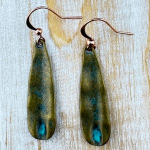 A pair of green earrings with blue accents, hanging from a curved hook on a wooden surface.