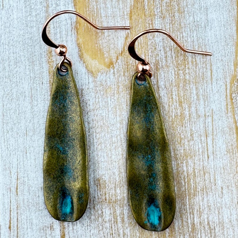 A pair of green earrings with blue accents, hanging from a curved hook on a wooden surface.