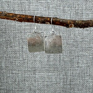 May include: A pair of silver square earrings with a hammered texture, hanging from a thin branch. The earrings have a simple, minimalist design and are suspended by silver hooks. The background is a textured gray fabric.