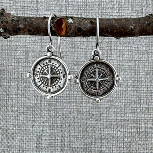 May include: Silver compass earrings with a detailed design. The earrings are hanging from a branch.