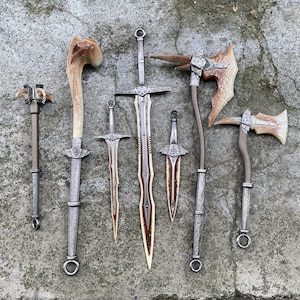 May include: An assortment of fantasy weapons, including swords, axes, and a mace, are displayed on a textured gray surface. The blades and heads have a metallic finish, with brown and cream-colored accents. The handles are brown and gray.