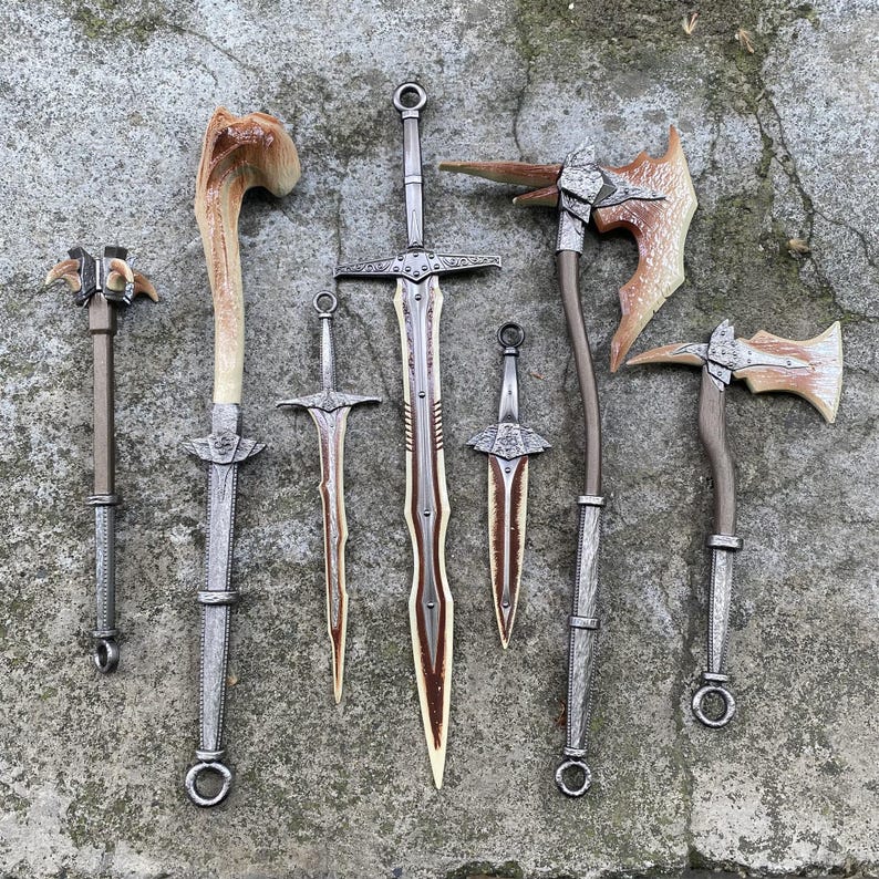 Puede incluir: Un surtido de armas de fantas&iacute;a, incluyendo espadas, hachas y una maza, se exhiben sobre una superficie gris texturizada. Las hojas y cabezas tienen un acabado met&aacute;lico, con detalles en marr&oacute;n y crema. Los mangos son marrones y grises.