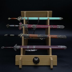 May include: Four decorative swords on a wooden stand. The swords have ornate handles and sheaths in teal, red, brown, and burgundy. The stand is made of light-coloured wood and features a decorative emblem. The swords are displayed on a black background.