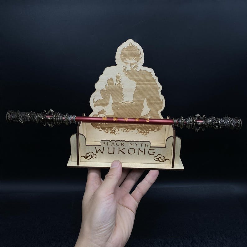 May include: Wooden stand with a laser-engraved image of a monkey holding a staff. The stand has the words "Black Myth Wukong" printed on it. A red and silver staff is resting on the stand.