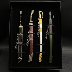 May include: A set of four miniature decorative swords displayed in a black box. The swords feature ornate handles and sheaths in various colors, including gold, red, white, and green. Each sword is secured with a black band and chain.