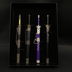 Op de afbeelding: Een set van vier decoratieve miniatuurzwaarden in een zwarte displaydoos. De zwaarden hebben sierlijke zilveren en gouden details, met zwarte, paarse en gouden handvatten. Eén zwaard heeft een ketting en een ander heeft een paarse kralenhandgreep.