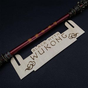 May include: A wooden display stand with the words "BLACK MYTH WUKONG" engraved on it. A red and gold staff with Chinese characters is resting on the stand.