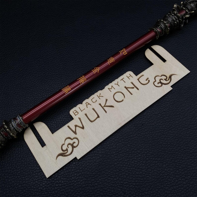 May include: A wooden display stand with the words "BLACK MYTH WUKONG" engraved on it. A red and gold staff with Chinese characters is resting on the stand.