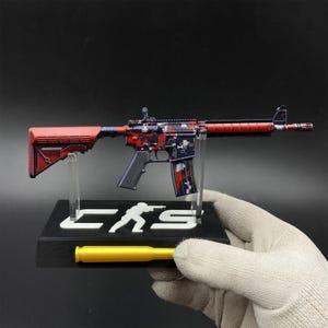 May include: A miniature rifle model featuring a red and black digital camouflage pattern on the receiver, with a solid red stock and barrel guard. It is mounted on a black display stand with the white text CKS and a yellow bullet replica.