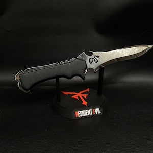 May include: A silver and black knife with a textured handle, displayed on a black stand. The stand features a red emblem and the text "RESIDENT EVIL". The knife has a curved blade and a decorative design.