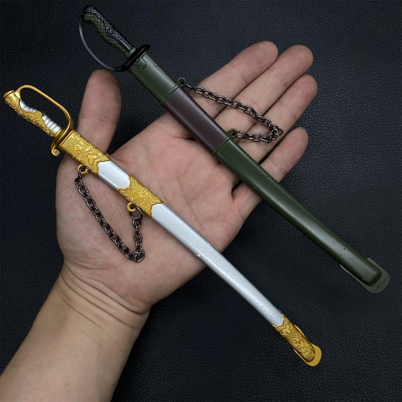 May include: Two decorative miniature swords. One sword has a silver blade with gold accents and a gold handle. The other sword is in a green sheath with a black handle. Both swords have a chain attached.