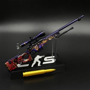 May include: A miniature sniper rifle model featuring a purple barrel and scope, decorated with a detailed graphic of a Japanese-style mask and red flame patterns. The model is mounted on a black display stand with a white silhouette and text.