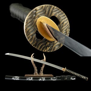Op de afbeelding: Een Japanse katana-zwaard met een zwarte handgreep en schede, met een zilveren lemmet. De pareerstang is goud en brons met een golfpatroon. Het zwaard rust op een houten standaard met geweivormige steunen, tegen een zwarte achtergrond.