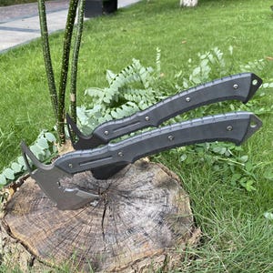 May include: Two tactical axes with matte grey metal heads and textured dark grey handles. Each axe features a curved blade, a pointed spike on the back, and a handle secured with three visible metal rivets, ending in a small circular lanyard hole.