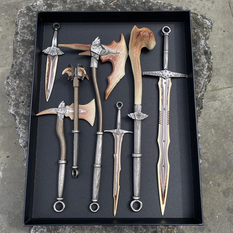 Puede incluir: Una colecci&oacute;n de armas de fantas&iacute;a, incluyendo espadas, hachas y una maza, dispuestas en una bandeja negra. Las hojas y cabezas tienen un acabado met&aacute;lico plateado, con mangos y detalles de madera. Las armas var&iacute;an en tama&ntilde;o y dise&ntilde;o.
