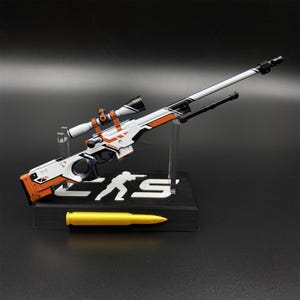 May include: A miniature sniper rifle model featuring a white, black, and orange color scheme with a mounted scope and bipod. The model rests on a black display stand with the letters CS and a white silhouette figure, accompanied by a yellow bullet.