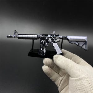 May include: A miniature model of an assault rifle featuring a black and white digital camouflage pattern on the receiver and barrel, a detachable magazine, a solid stock, and a front sight post, mounted on a black plastic display stand.