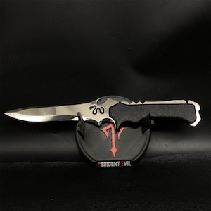May include: A silver-colored dagger with a black handle rests on a black stand. The stand features a red and black emblem and the text "RESIDENT EVIL". The dagger has a curved blade and a decorative design near the handle.