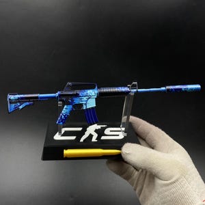 May include: A miniature rifle model featuring a blue and black splatter paint finish, mounted on a black display stand with the white text CKS and a silhouette graphic. A yellow bullet-shaped object is attached to the front of the base.