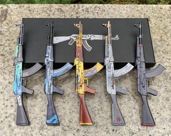 May include: Five miniature rifle models arranged in a row on a black rectangular box. Each model features a distinct finish, including blue, grey, gold, patterned, and black designs, with a white line drawing of a rifle printed on the box.