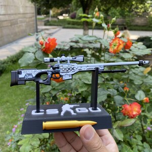 May include: A detailed model of a sniper rifle, predominantly light gray with black and white accents. The rifle features a scope and is mounted on a black base with the text "CS GO" and a golden bullet. The background includes vibrant orange flowers.