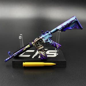 May include: A miniature rifle model featuring a vibrant purple and blue cosmic pattern with a prominent astronaut helmet graphic on the receiver. The model is displayed on a black stand with a single gold-colored bullet replica positioned in front.