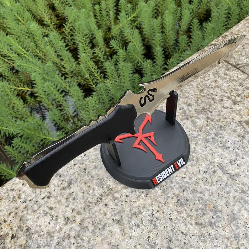 May include: A silver-colored knife with a black handle, displayed on a black stand. The stand features a red emblem and the text "RESIDENT EVIL". The knife has a serrated edge and a decorative design.