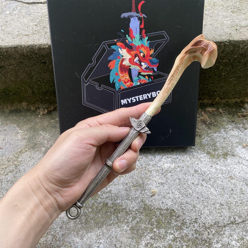 Puede incluir: Un abrecartas artesanal &uacute;nico, con un mango con forma de hueso y una hoja plateada. El mango tiene un dise&ntilde;o curvado con tonos marrones y beige. La hoja presenta un elemento decorativo en forma de cruz. El fondo incluye una caja negra con una ilustraci&oacute;n de drag&oacute;n.