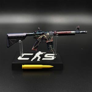 May include: A miniature rifle model featuring a dark, multi-colored graphic finish with intricate patterns, mounted on a black display stand with the white text "CS" and a silhouette figure. A single yellow bullet replica rests in front of the base.