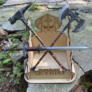 May include: A collection of fantasy weapons, including a hammer, axe, and sword, displayed in a wooden stand. The stand features the text "SKYRIM" and a decorative emblem. The weapons have intricate designs and metallic finishes.