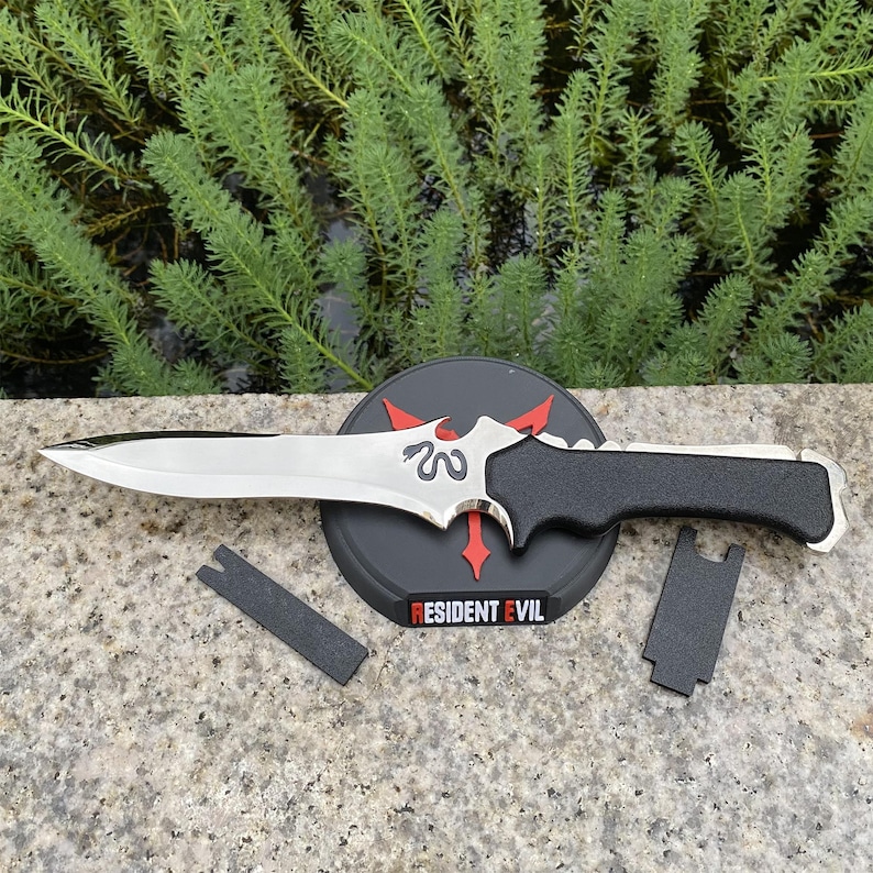 May include: A silver and black dagger with a black handle, resting on a black stand with the text "RESIDENT EVIL" in red and white. The dagger has a decorative design on the blade.
