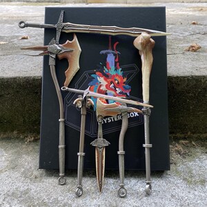 Puede incluir: Una colecci&oacute;n de armas de fantas&iacute;a en miniatura, que incluye espadas, hachas y un objeto con forma de hueso, dispuestas alrededor de una caja negra con una ilustraci&oacute;n de drag&oacute;n colorido. Las armas tienen tonos met&aacute;licos y marrones.