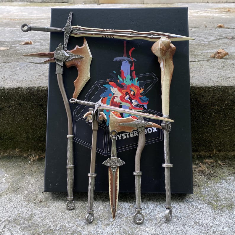 Puede incluir: Una colecci&oacute;n de armas de fantas&iacute;a en miniatura, que incluye espadas, hachas y un objeto con forma de hueso, dispuestas alrededor de una caja negra con una ilustraci&oacute;n de drag&oacute;n colorido. Las armas tienen tonos met&aacute;licos y marrones.
