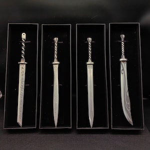 Puede incluir: Cuatro espadas decorativas en miniatura con hojas plateadas y empuñaduras retorcidas se exhiben en cajas negras individuales. Las hojas presentan diseños grabados, incluyendo patrones geométricos y motivos estilizados. Las espadas se presentan sobre un fondo oscuro.