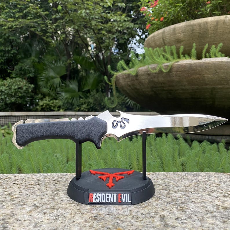 May include: A silver and black knife with a curved blade, displayed on a black stand. The stand features a red emblem and the text "RESIDENT EVIL". The knife has a black handle and a silver blade with a decorative design.