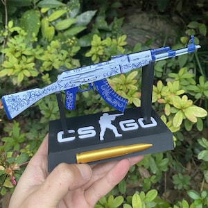 以下が含まれることがあります： 青と白の花柄模様の装飾用AK-47モデル。黒い台座に「CS GO」の文字が記されています。金色の弾丸が台座の前に置かれています。緑の葉の背景の前にモデルが保持されています。