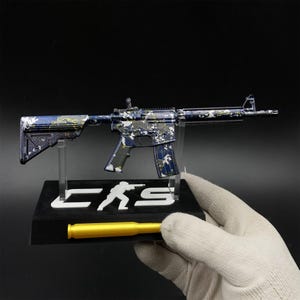 May include: A miniature rifle model featuring a blue, white, and yellow camouflage pattern. The model is mounted on a black display stand with the white text CKS and a silhouette figure, accompanied by a single gold-coloured bullet replica.