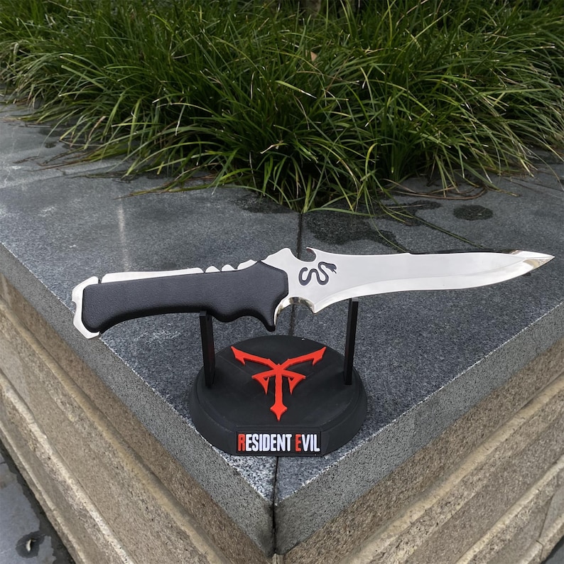 May include: A silver and black dagger with a serrated handle rests on a black stand. The stand features a red emblem and the text "RESIDENT EVIL". The dagger has a curved blade and a decorative design near the handle.