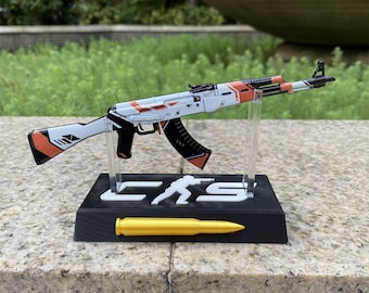 May include: A miniature rifle model featuring a white, orange, and black geometric pattern, mounted on a black rectangular stand with the text CS and a silhouette figure. A single gold-colored bullet replica is attached to the front of the base.