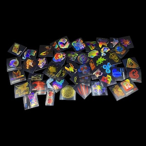 Könnte beinhalten: Eine Sammlung holografischer Aufkleber mit verschiedenen Designs, darunter ein Totenkopf, ein Spinnennetz und eine Krone. Einige Aufkleber tragen Texte wie "KATOWICE 2014" und "WELCOME TO THE CLUTCH". Die Aufkleber befinden sich auf schwarzem Hintergrund.