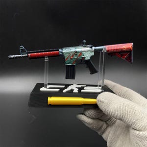 May include: A miniature rifle model featuring a teal and red diagonal striped pattern on the receiver, a black magazine, and a red stock. The model is mounted on a black display stand with white lettering and a yellow bullet-shaped accent piece.