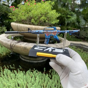 May include: A miniature rifle model featuring a vibrant blue and red graphic pattern on the receiver and barrel. The model is mounted on a black display stand with white lettering reading CKS and a yellow cylindrical accent piece at the base.