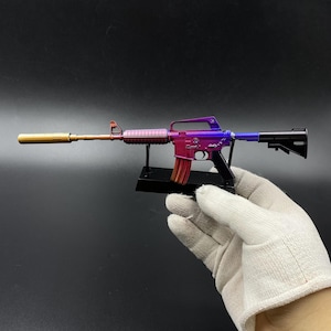 May include: A miniature model of an assault rifle featuring a metallic gradient finish that transitions from gold at the barrel to purple and pink on the receiver. The model includes a black stock, a detachable magazine, and a black display stand.