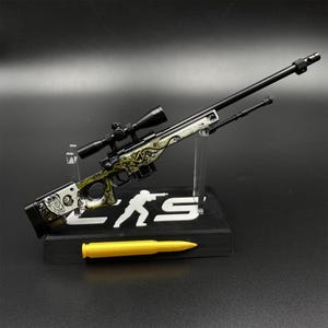Könnte beinhalten: Ein Miniatur-Scharfschützengewehr-Modell mit schwarzem Lauf und Zielfernrohr, dessen Schaft mit gelb-schwarzen Krakenmustern und einer Totenkopf-Grafik verziert ist. Es steht auf einem schwarzen Ständer mit weißer Silhouette und einem gelben Modellpatron.