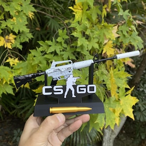 May include: A miniature replica of a rifle, featuring a white and black design, rests on a black stand with the text "CS GO". A golden bullet is positioned below. The background includes green and yellow leaves.
